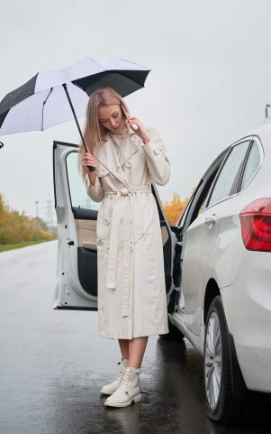 Beautiful woman standing on a road near her stopped white car with punctured car tire. Female driver calling car service for help, holding umbrella.